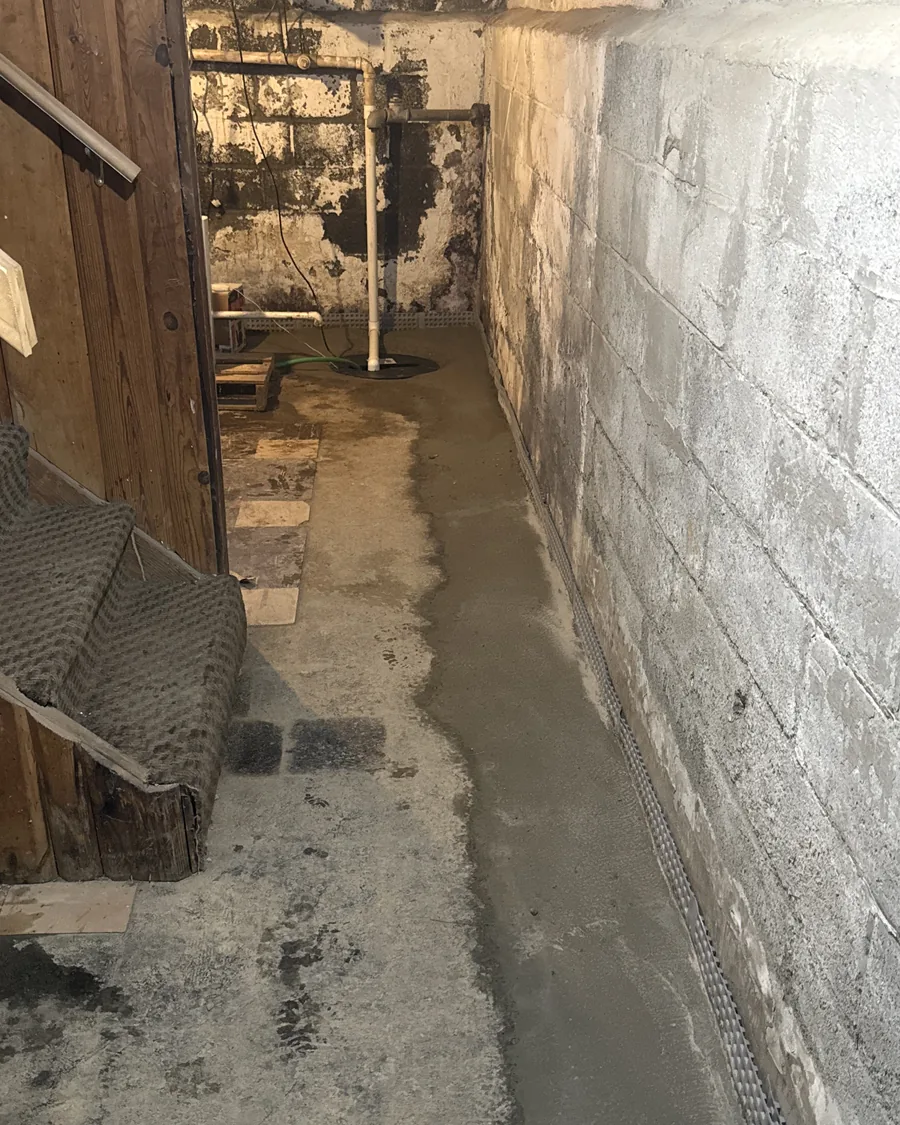 Perimeter drainage track and fresh concrete running along a basement wall toward the sump