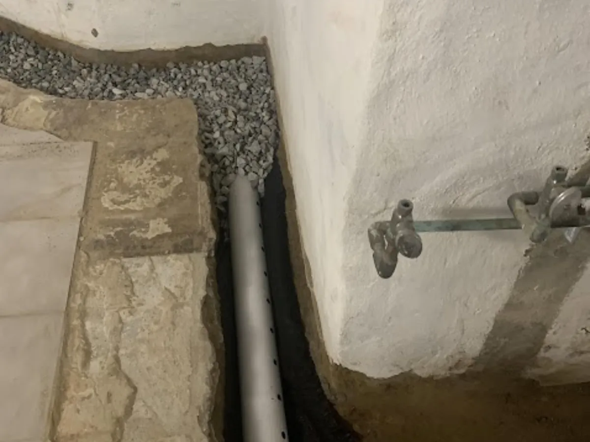 Perforated drainage pipe laid on black filter fabric in a trench beside a stone foundation, surrounded by washed stone