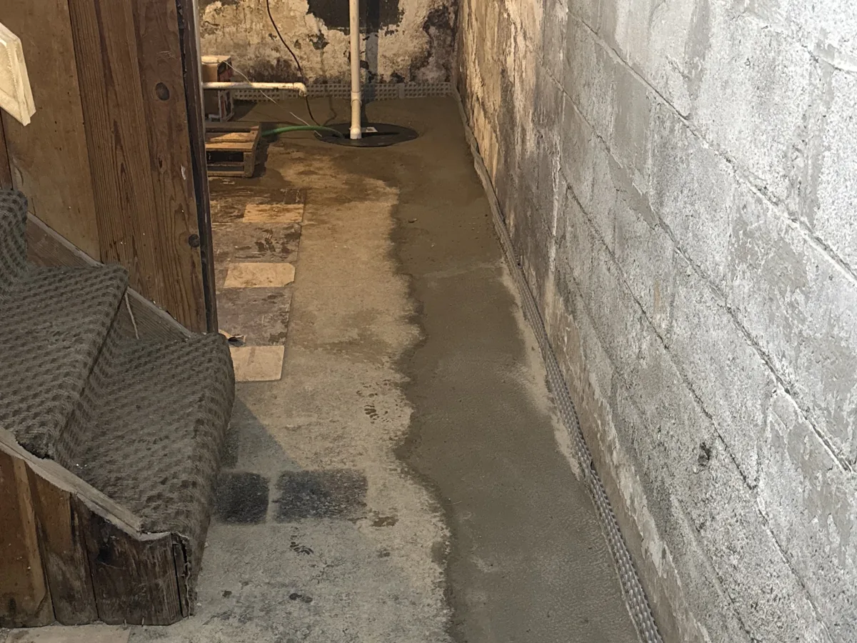 Perimeter drainage track and freshly poured concrete running along a basement wall toward a sump