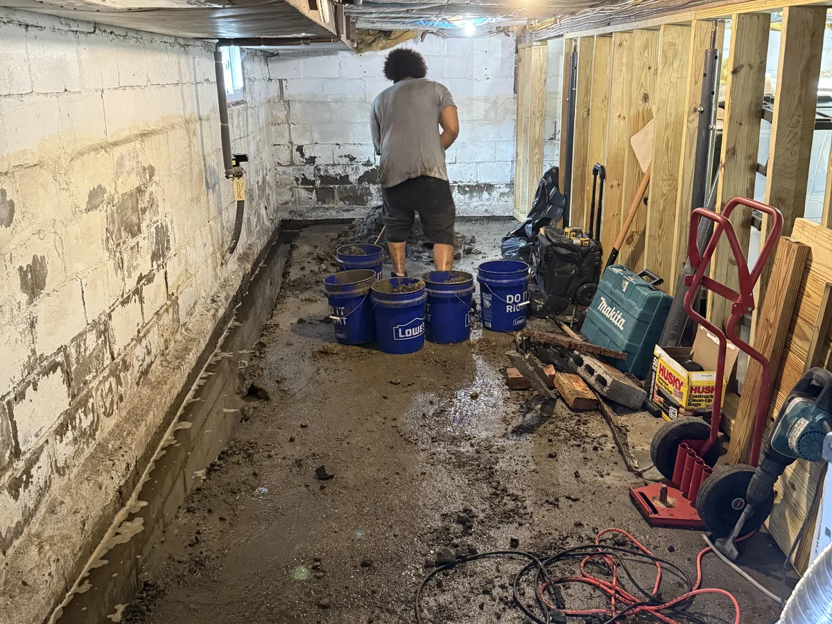 Crew member excavating an interior perimeter trench beside a block foundation wall, with buckets of spoil